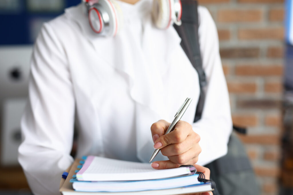 woman in a white shirt with headphones on her neck holds notebooks and pen